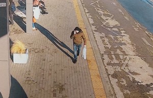Rozmawiający przez telefon mężczyzna, ubrany w brązową kurtkę, spodnie typu jeans, czarne buty i czapkę. W lewej ręce niesie reklamówkę