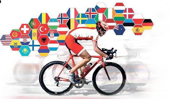 Na tle flag narodowych różnych państw umieszczony kolarz, ubrany w strój splotowy w kolorze biało czerwonym podczas jazdy na rowerze