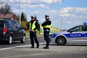 Dwóch policjantów stojących przy ulicy, przed radiowozem, a obok nich przejeżdża samochód. Pora dzienna