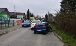 Cztery samochody uczestniczące w kolizji. Na chodniku widoczni świadkowie i uczestnicy zdarzenia. W tle pojazdy służb ratowniczych