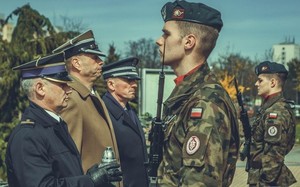 Delegacja przedstawicieli służb mundurowych podczas rocznicowych uroczystości. Skład delegacji stanowi strażak trzymający w ręku znicz, żołnierz z okolicznościową wiązanką kwiatów oraz policjant w umundurowaniu wyjściowym