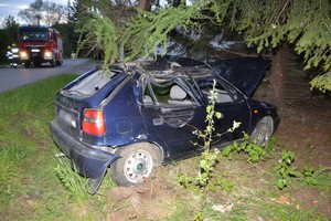 Uszkodzenia powypadkowe skody. Samochód poza drogą, oparty o drzewo. W tle wóz strażacki z włączonymi sygnałami świetlnymi oraz strażacy