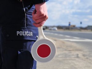 Trzymana w ręku funkcjonariusza tarcza do zatrzymywania pojazdów na tle napisu policja na kieszeni spodni służbowych. W tle ulica