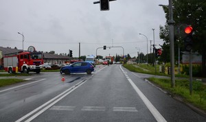 Uszkodzenia przedniej części peugeota stojącego w poprzek skrzyżowania. W tle pojazdy służb ratowniczych