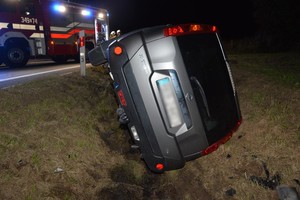 Leżący na boku w przydrożnym rowie nissan. Na dalszym planie wóz strażacki