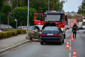 Uszkodzenia powypadkowe pojazdów, na dalszym planie samochód strażacki. Obok ustawione na jezdni pachołki drogowe