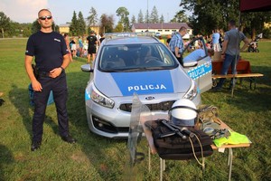 Policjant stojący obok radiowozu podczas pikniku integracyjnego. W tle uczestnicy wydarzenia