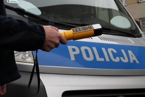 policjant trzymający w ręku urządzenie do badania stanu trzeźwości, na dalszym planie radiowóz