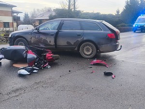 Uszkodzenia powypadkowe samochodu, obok leżący na jezdni uszkodzony motocykl. W tle radiowóz z włączonymi światłami błyskowymi